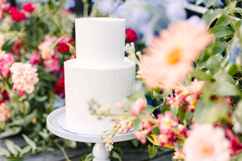 Two tier white wedding cake on stand surrounded by bright flowers. 