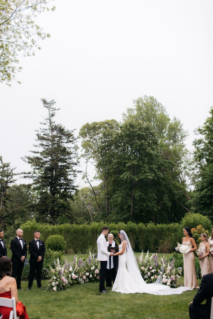 Bride and groom hold hands while exchanging vows in front of floral semi-circle showing one of the 2025 wedding floral trend.