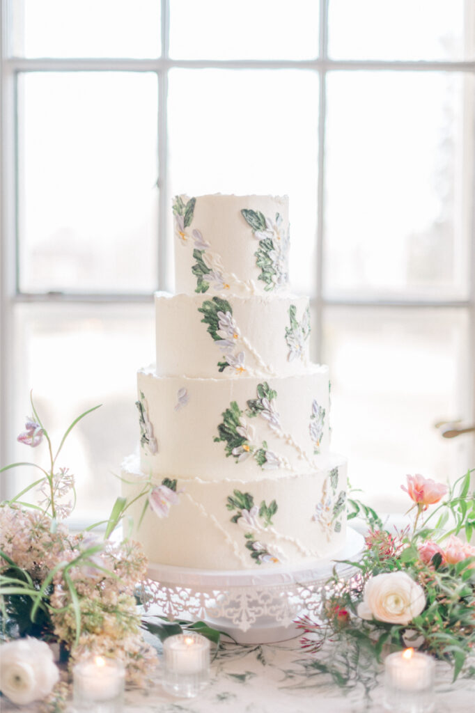 White four tier wedding cake with pressed edible flowers, surrounded with table of flowers. 