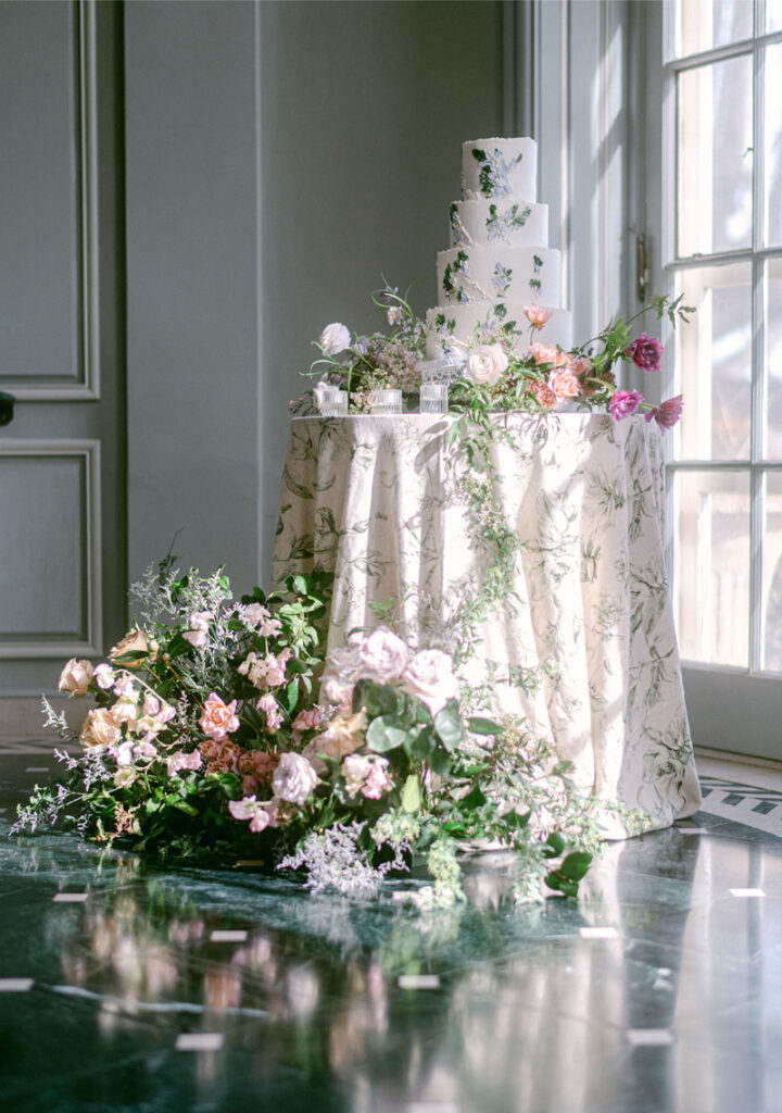 Four tier white wedding cake with edible pressed flowers surrounded by a cake garden of flowers with a large flower arrangement sitting on floor in front of cake table. 