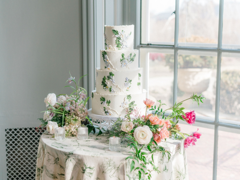 Four tier white wedding cake with edible pressed flowers surrounded by a cake garden of flowers, showing one of the 2025 wedding floral trends.