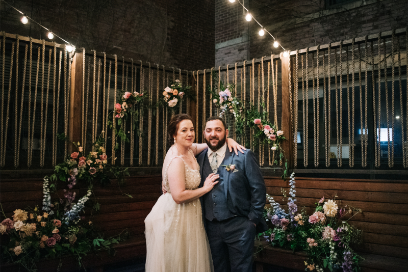 Bride and groom stand in front of untraditional ceremony arch made of flowers.