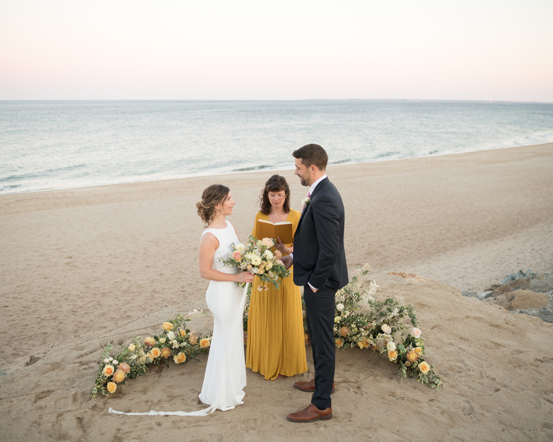 Couple exchanging vows on New England beach with a floral altar surrounding them. 