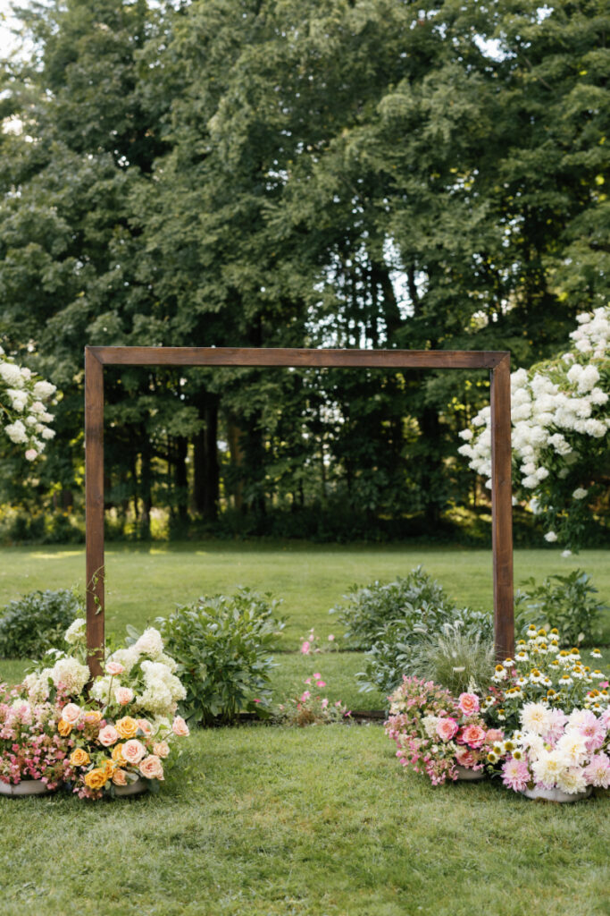 Wooden rectangular wedding arch with large summer flower arrangements at the base. 