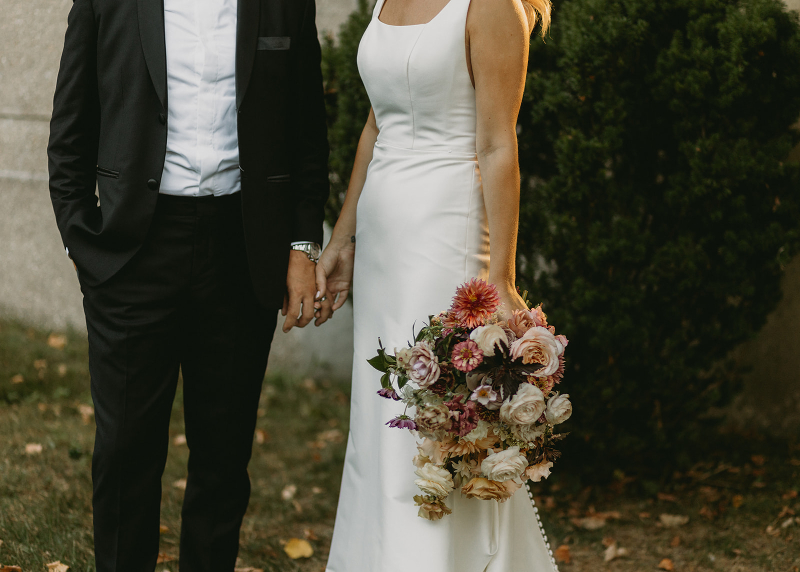 Bride and groom hold hands as they pose with bouquet of seasonal flowers.