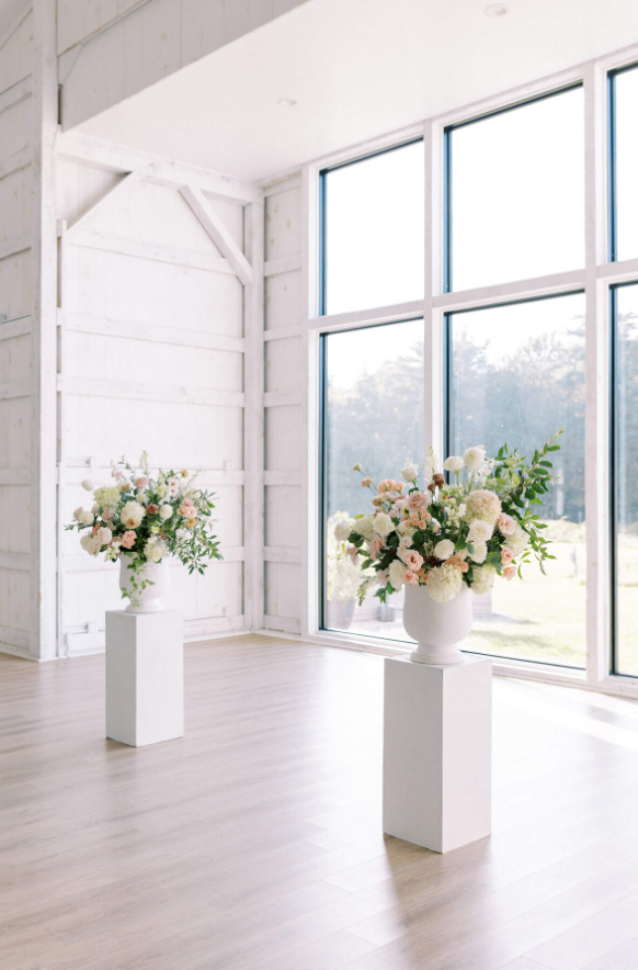 Elegant floral arrangements in white urns with blush, ivory, and green blooms sit atop white pedestals in a bright, modern venue.