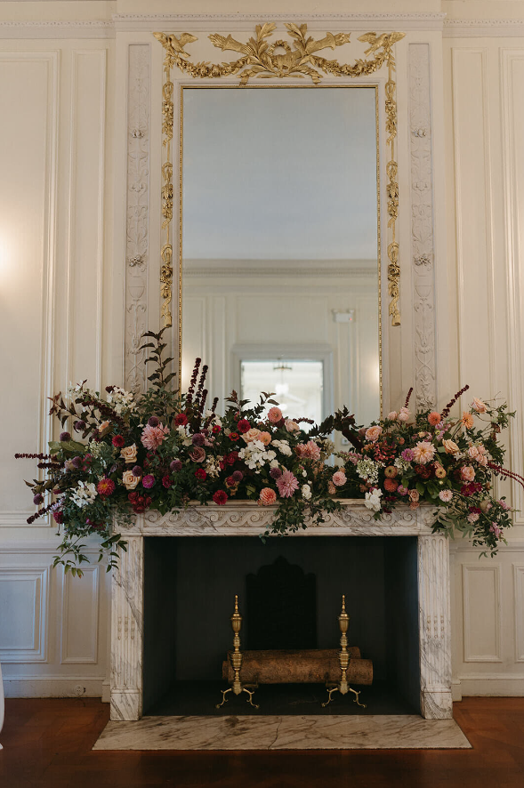 An opulent mantel adorned with a vibrant floral display of pink, burgundy, and greenery in an elegant room with a gold mirror.