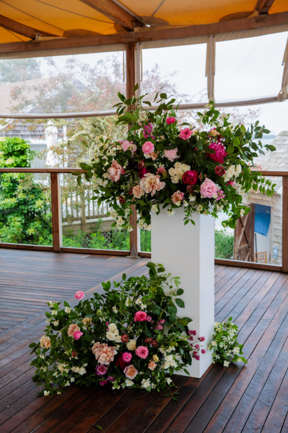 A vibrant floral arrangement with lush greenery and pink, white, and magenta blooms displayed on a white column and at its base on a wooden deck.