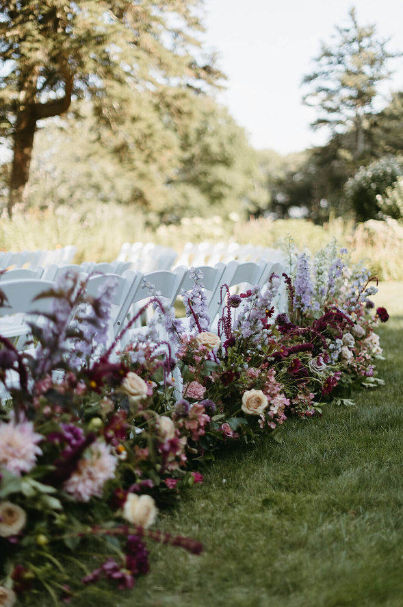 A lush aisle arrangement featuring soft lavender, deep burgundy, and blush blooms set against a green outdoor ceremony backdrop with white chairs.