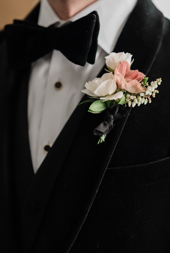 A classic boutonniere featuring a white rose, blush accent bloom, delicate greenery, and lily of the valley, tied with a black ribbon.