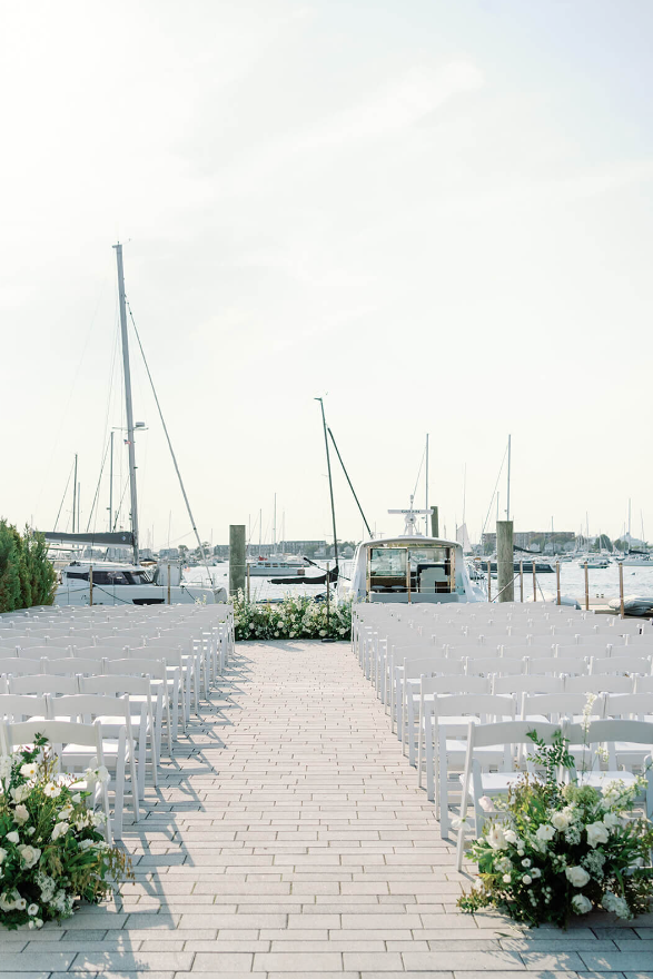 waterfront ceremony setup featuring white chairs, lush floral aisle arrangements, and a view of sailboats in the background.