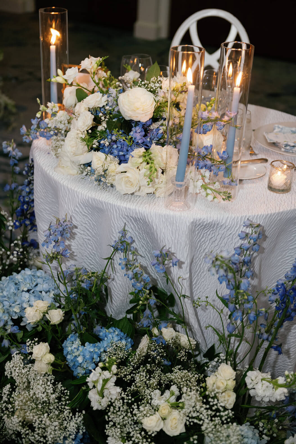 A sweetheart table surrounded by cascading blue and white florals, and greenery accented with blue taper candles | wedding floral design budget