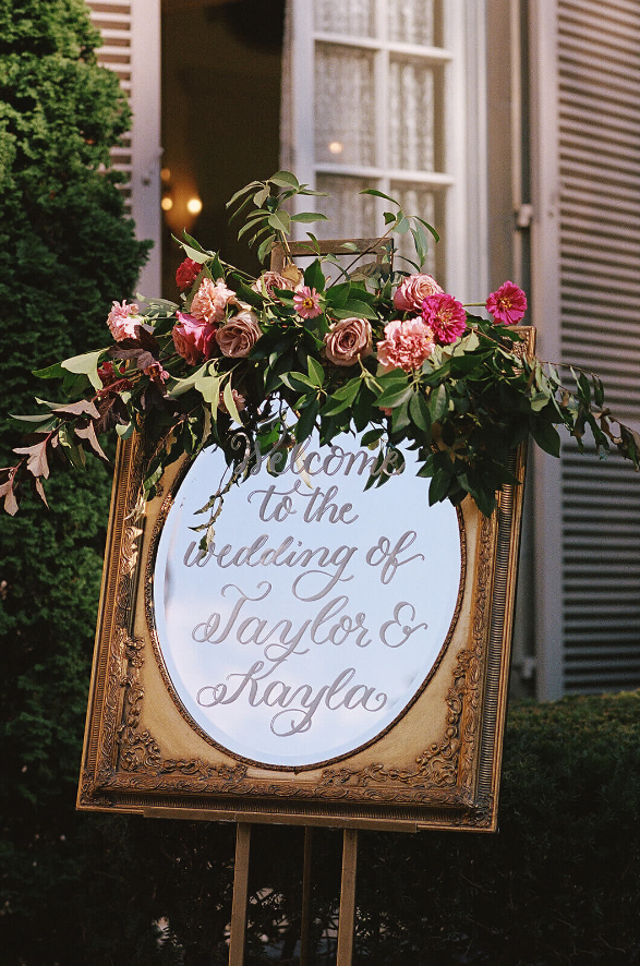 A wedding welcome sign framed by an elegant arrangement of greenery and burgundy, blush, and pink florals | wedding floral design budger