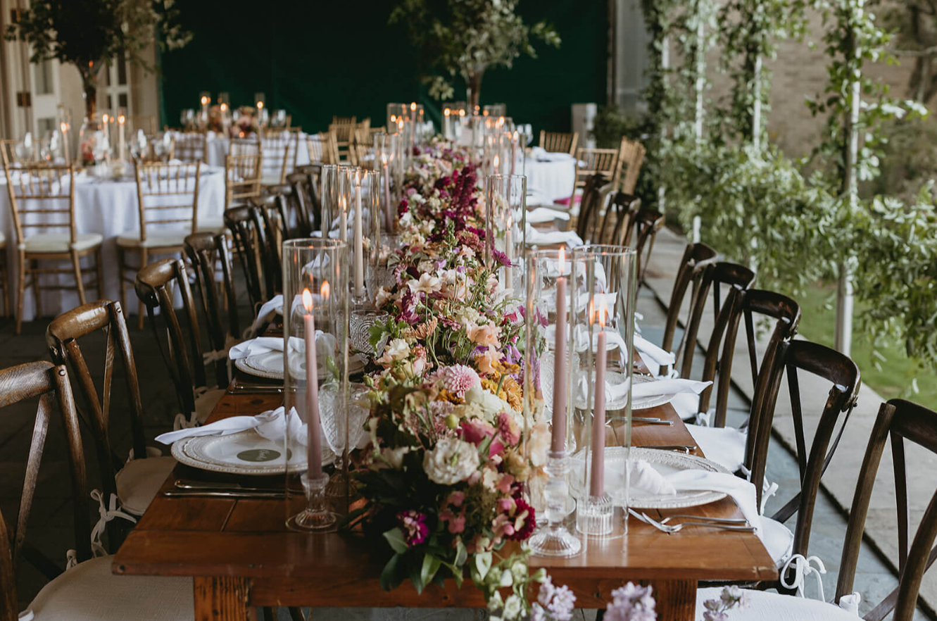 A long reception table with a lush floral garland in pink, ivory, and burgundy tones with taper candles