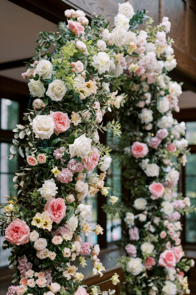 A floral arch features an abundance of roses, peonies, and greenery in soft pastel shades of white, blush pink, and peach.